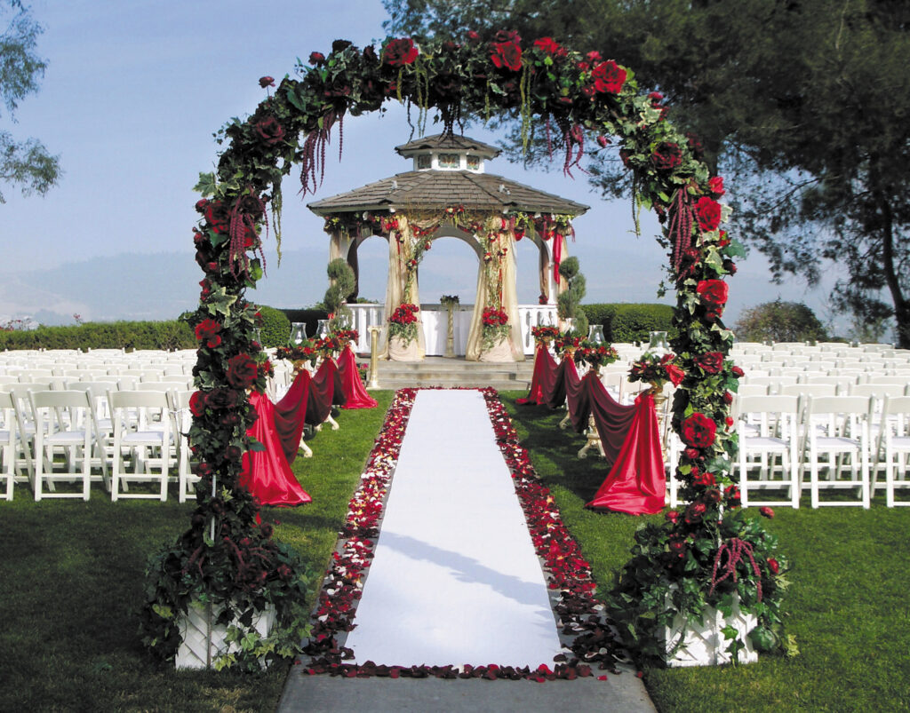 Elegant outdoor wedding ceremony setup with a white aisle, red rose petals, floral arch, and decorated gazebo surrounded by white chairs